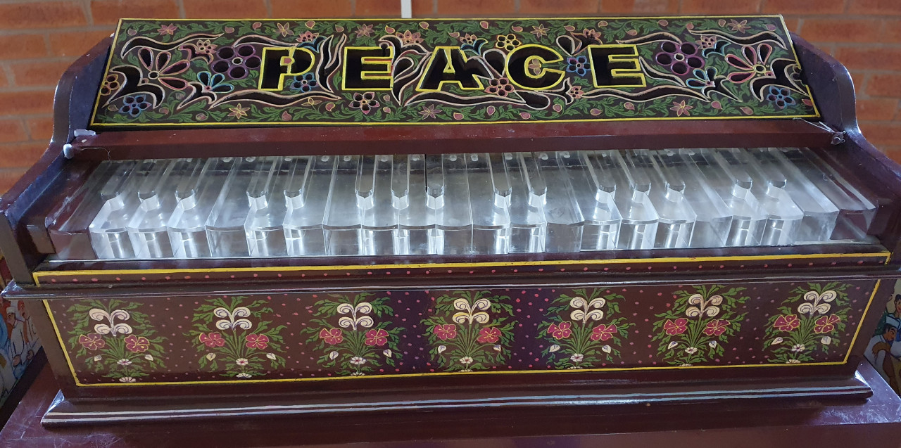 Image shows small, ornate keyboard with clear plastic keys and a polished lacquered wooden frame with flowers. The word Peace is written in black with a bright yellow outline. In the background is red brickwork.